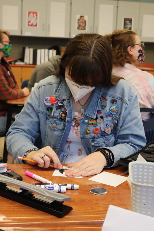 Student wearing jean jacket decorates pronoun pin using markers and paper.